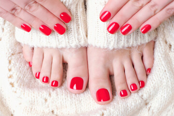 Female feet and hand with red pedicure and manicure on white knitted surface, top view.