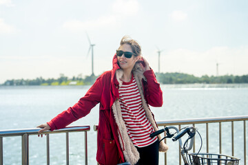 Obraz premium Close up Woman with red coat and sun glasses lean to fence and touch coat hood also hold hand of bicycle stand in front of wind turbines or windmill near water reservoir.