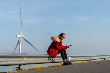 Wide shot of Caucasian woman with red coat and sunglasses sit on barrier of roadside and use mobile phone on mountain near windmill or wind turbine with morning light.