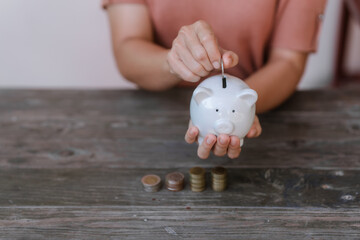 hand placing a coin into a white piggy bank on a table against a pale pink background.