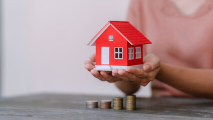  person holding a small red model house above a row of increasing coin stacks, symbolizing real...