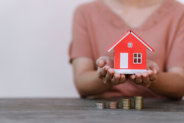  person holding a small red model house above a row of increasing coin stacks, symbolizing real estate investment or savings.