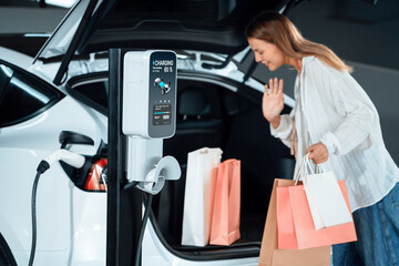 Young woman travel with EV electric car to shopping center parking lot charging in downtown city...
