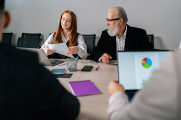 View from shoulder to female trainee reporting new strategy to mature business partner. Motivated skilled young businesswoman having conversation with corporate male mentor sitting at office desk.