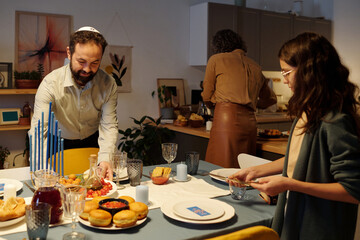 Cute girl and her father putting cutlery and homemade food on table while mature woman preparing something by kitchen table