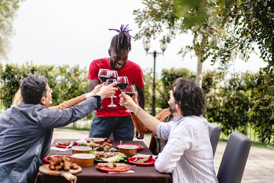 Diverse group of friends laughing and toasting with red wine at an outdoor table filled with delicious food.