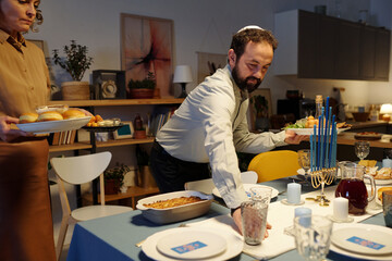Bearded Jewish man in yarmulke helping his wife serve festive table with homemade food and drinks while waiting for guests