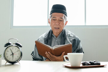 Elderly Asian muslim man reading holy book of Quran or Koran while sitting at dining table waiting...