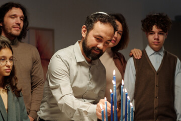 Mature man in Jewish skullcap putting burning blue candle on menorah candlestick while celebrating Hanukkah at home with his family