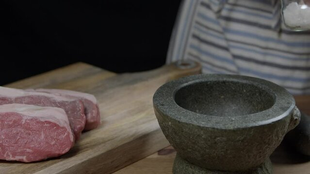 Hands depositing salt in the mortar and then crushes it to sprinkle on the cut of meat