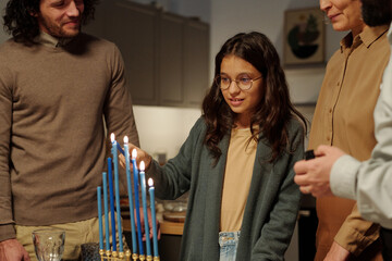 Pretty pre-teen girl lighting blue candles of menorah while standing among her family and preparing...
