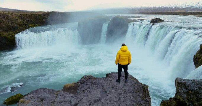 View Of A Man Standing On A Cliff Next To Godafoss Waterfall In Iceland - Aerial Pullback