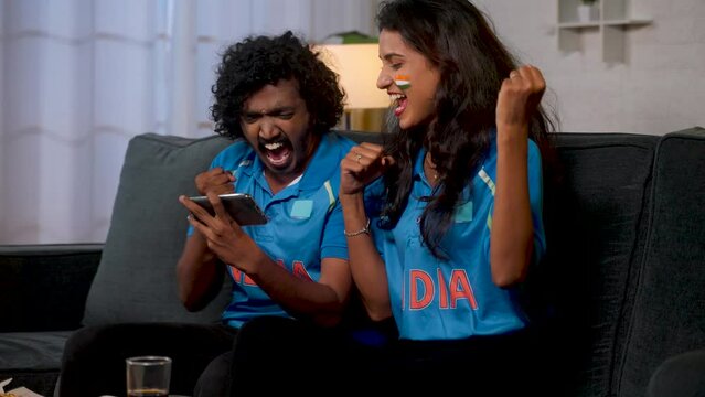 excited young friends in Indian jersey celebrating victory of team indias in cricket while watching on mobile phone at home - concept of tournament, sports match and supporters