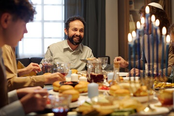 Cheerful mature man looking at one of guests during Hanukkah dinner with his family while enjoying...