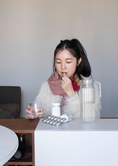 Asian girl taking medicines, pills, supplements at home, in winter clothing and scarf, flu, cold