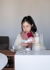 Asian girl taking medicines, pills, supplements at home, in winter clothing and scarf, flu, cold