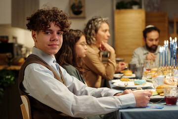 Youthful Jewish boy in casualwear looking at camera by dinner table served with homemade food and drinks against members of his family