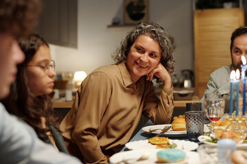 Happy mature Jewish woman looking at one of her children during chat while sitting by table served...