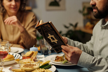 Bearded Jewish man reading text from Torah by served table while sitting in front of his family...