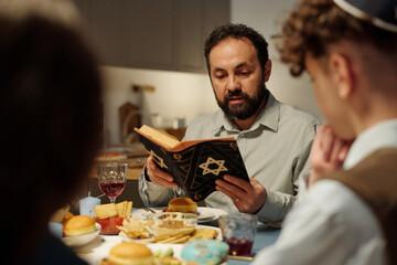 Focus on bearded rabbi and head of Jewish family reading text from Torah while sitting by served table in front of his two children