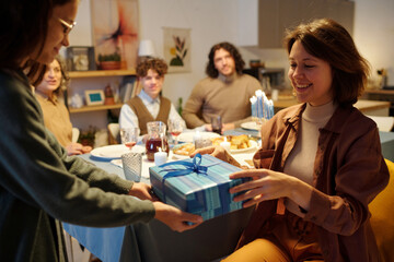 Young smiling woman taking blue giftbox with Hanukkah present from hands of her daughter against other members of family having dinner