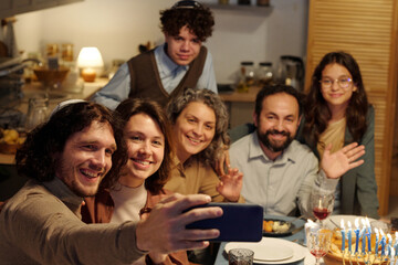 Happy young Jewish man with smartphone taking selfie of large family or communicating in video chat during Hanukkah dinner at home