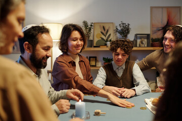 Young smiling woman putting coin on center of table while betting during leisure game with members of her family after Hanukkah dinner