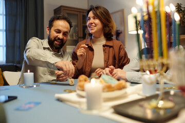 Happy mature man spinning dreidel on table while sitting next to cheerful young woman looking aside during leisure game after dinner