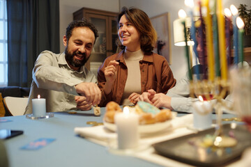 Happy mature man spinning dreidel on table while sitting next to cheerful young woman looking aside...