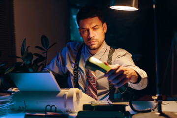 Young serious businessman with phone receiver in hand sitting by workplace in dark office lit by...