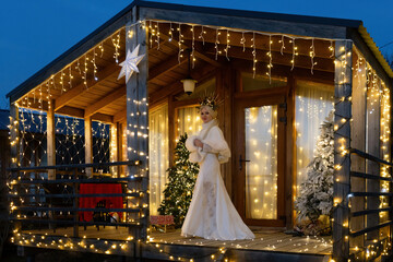 Happy blonde as Snow Maiden in white costume. Wooden house exterior Festive setting. Character with crown creating festive scene.