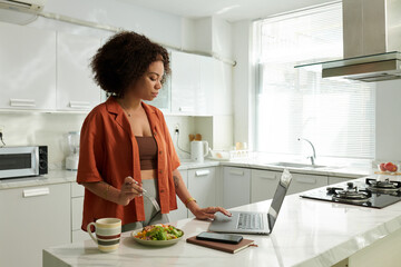 Serious woman eating vegetable salad for breakfast when uploading files on website