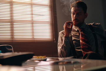 Young serious businessman in formalwear smoking cigarette while sitting by workplace in dark office...
