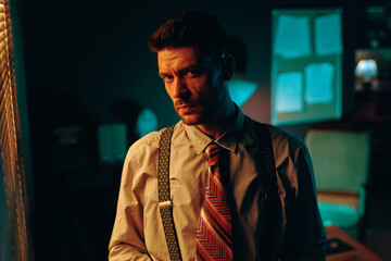 Young serious man in shirt, tie and suspenders looking at camera while standing in dark office with window covered with venetian blinds
