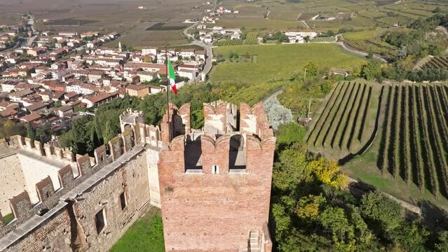 Orbiting On Restored Soave Castle Tower With Italian Flag. Vinery Fields In The Background In Soave Verona, Italy. Aerial Shot