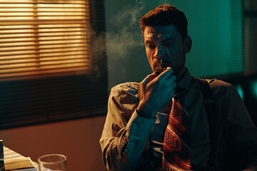 Pensive young man with tobacco pipe in mouth sitting by workplace in front of camera during work in dark office with blinds on window