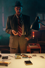 Young elegant man in old-fashioned suit and hat looking at camera while standing by table with set of photos and notepad in dark office