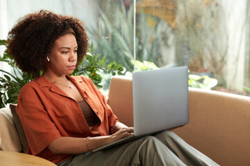 Serious Hispanic businesswoman sitting on comfy couch at home and reading reports on laptop