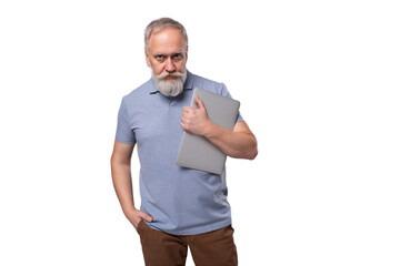 a gray-haired mature man with a beard and mustache in a t-shirt and trousers holds a laptop