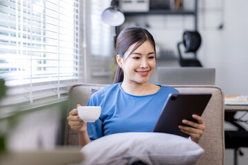 Asian young woman sitting on the yellow sofa using digital tablet and coffee cup and learning...