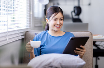 Asian young woman sitting on the yellow sofa using digital tablet and coffee cup and learning shopping or working online near window