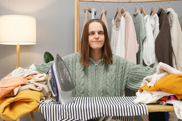 Confused angry brown haired woman wearing knitted shirt ironing clothing while sitting in her...