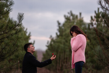 Fototapeta na wymiar Young man in black coat kneels in front of beautiful woman, proposes marriage in forest