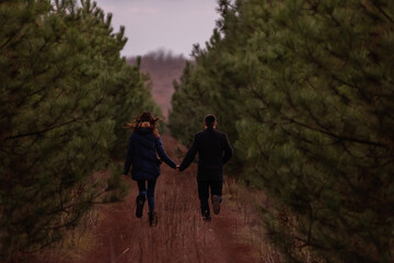 Happy couple running through forest holding hands. Man in coat and a young woman are fooling around