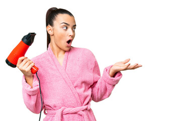 Young woman in a bathrobe holding a hairdryer over isolated chroma key background with surprise...