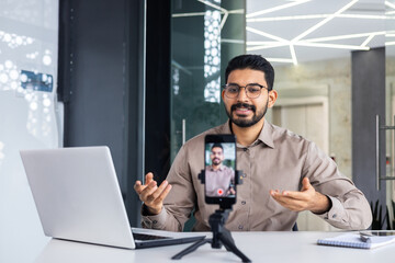 Successful business trainer teacher recording online training video course, man inside office at workplace using phone and tripod, sitting at desk with laptop