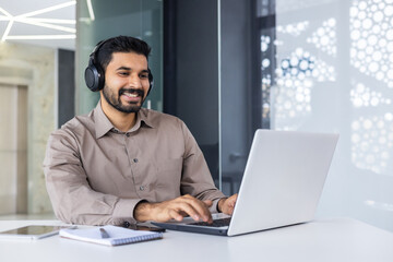Young successful Indian programmer at workplace inside office, man with headphones smiling...