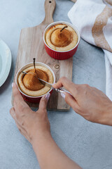 A pear soufflé in a red bowl stands on a wooden board, women's hands cut the souffle with vintage cutlery, next to it is a kitchen towel.