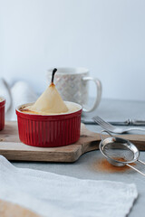 pear soufflé in a red bowl stands on a wooden board, behind is a white cup of coffee, next to it is a sieve with cinnamon and a kitchen towel
