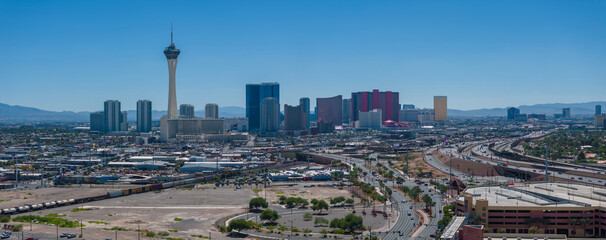 Aerial view of Las Vegas, Nevada, highlighting the Stratosphere Tower amid a bustling cityscape with green spaces, under a clear blue sky with mountains beyond.